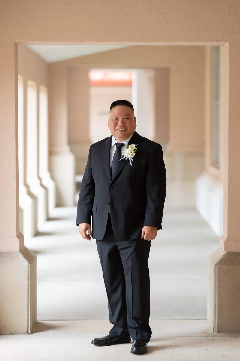 Groom Ronald stands confidently in a charcoal suit with white boutonniere in an elegant hallway with dramatic lighting and architectural columns.