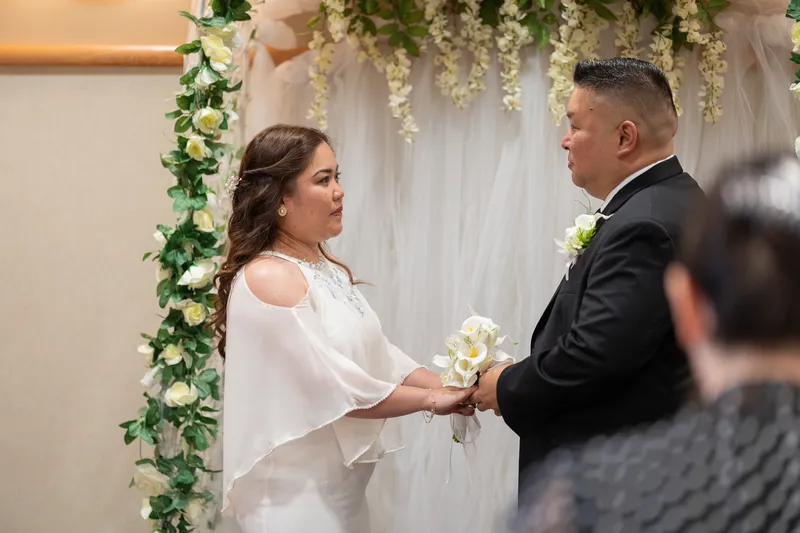 Ronald and Vanessa exchange vows during their wedding ceremony, holding hands beneath a beautiful floral arch adorned with white roses and greenery.