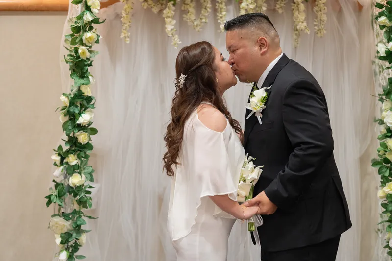 Ronald and Vanessa share their first kiss as newlyweds under a beautiful floral arch decorated with white roses and greenery.