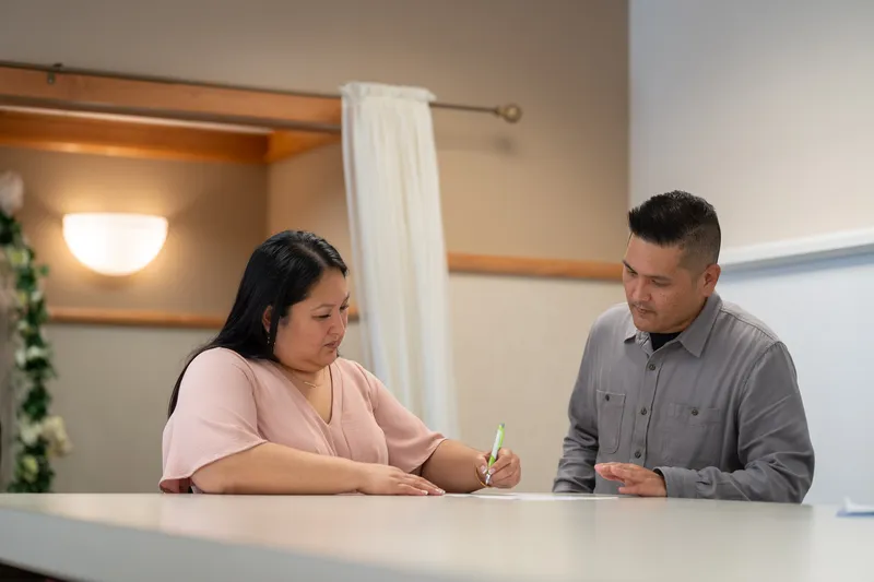 Ronald and Vanessa sit together at a white table during what appears to be their engagement or wedding planning session, with Vanessa writing while Ronald watches attentively.