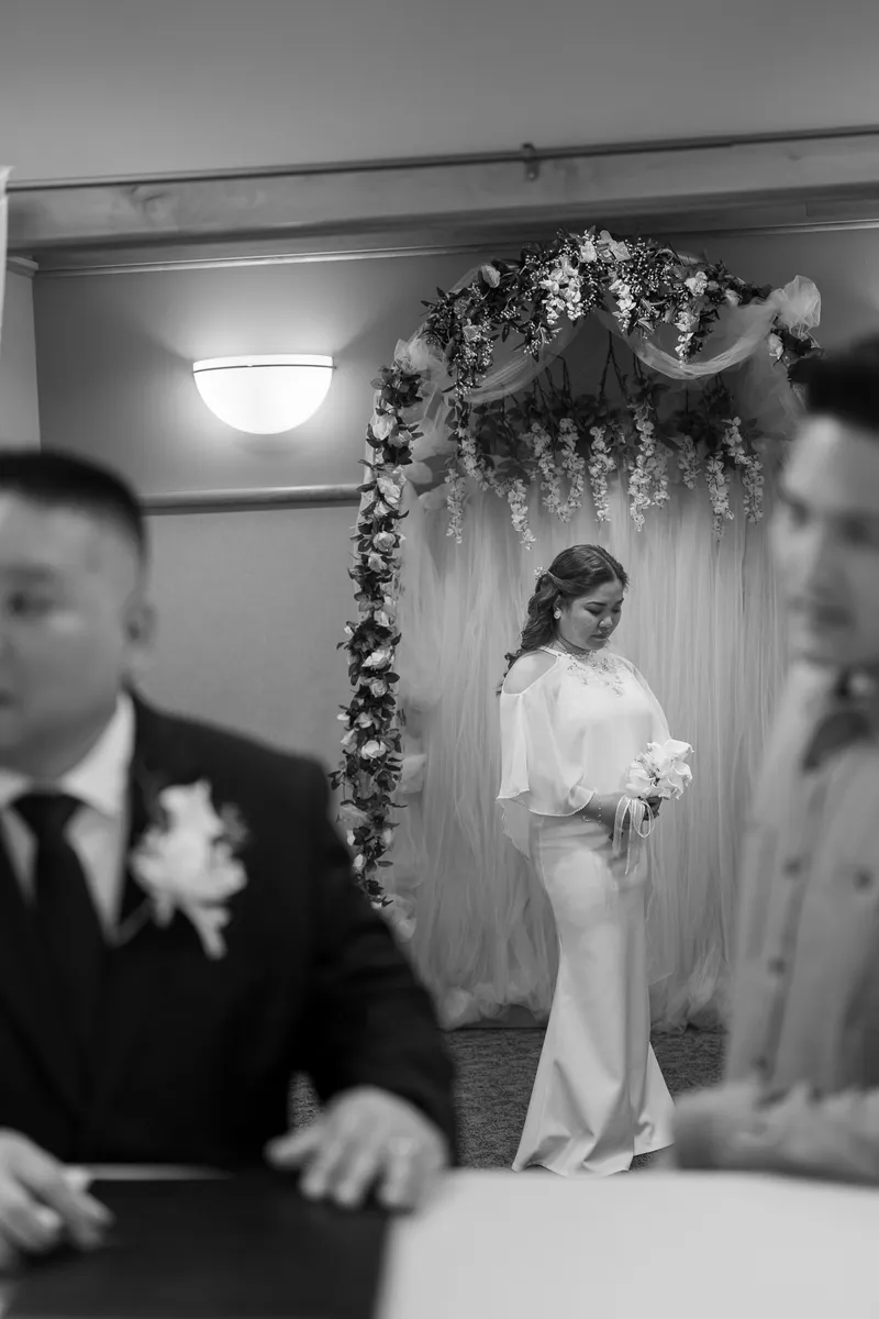 The bride walks down the aisle in her elegant wedding dress, passing under a beautiful floral arch while guests look on during the ceremony.