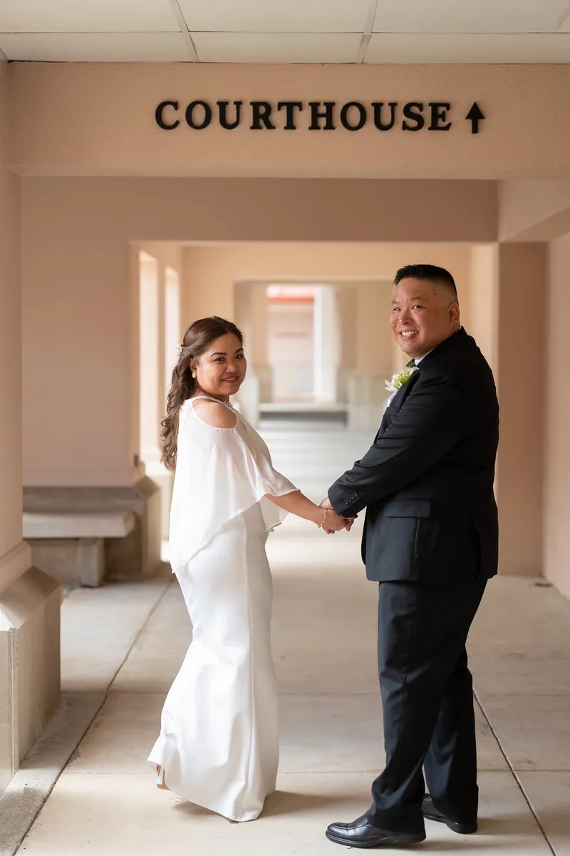 Ronald and Vanessa hold hands and smile at the camera in a courthouse hallway, celebrating their intimate wedding ceremony.