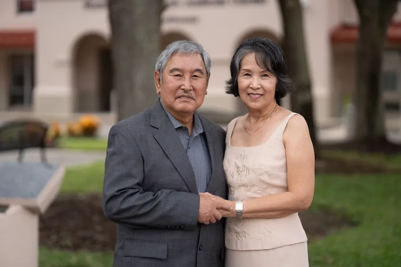 Ronald and Vanessa pose together elegantly dressed in front of a beautiful building with manicured landscaping.