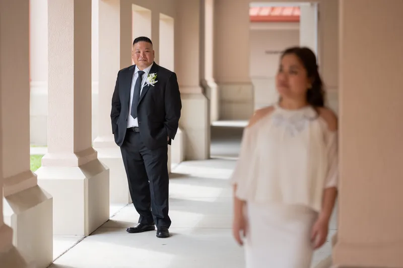Ronald in a classic black tuxedo walks down a columned corridor while Vanessa in flowing white appears softly blurred in the foreground, creating an intimate first look moment.
