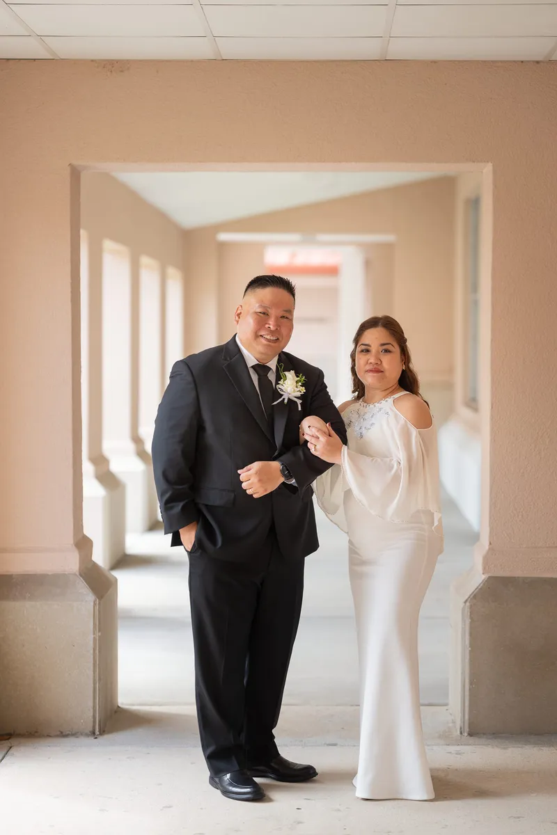 Ronald and Vanessa stand together in a courthouse hallway, he in a dark suit with boutonniere and she in a flowing white