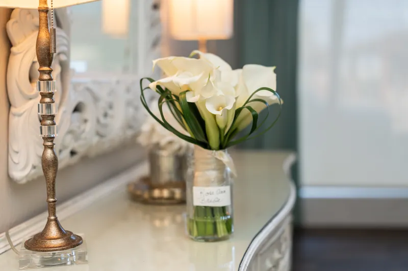 White calla lilies in a clear vase with green stems sit on a white surface near a gold bracelet.