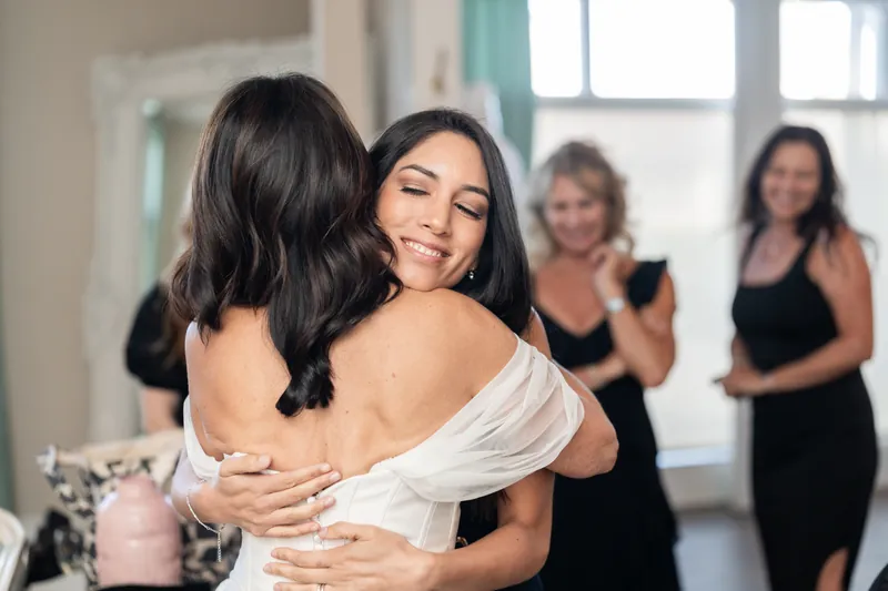 Rose Ann hugs a woman from behind while smiling, with guests in black dresses watching at The White Room.