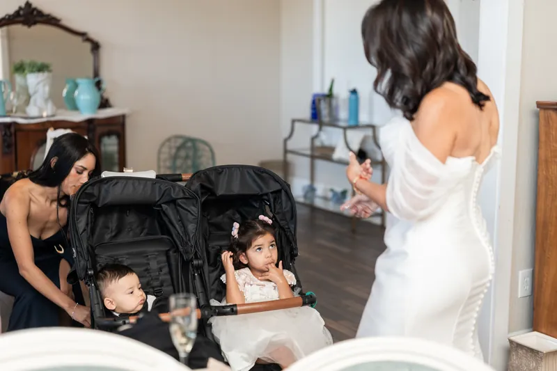 Rose Ann in her white wedding dress bends down to interact with young children in a stroller at The White Room.