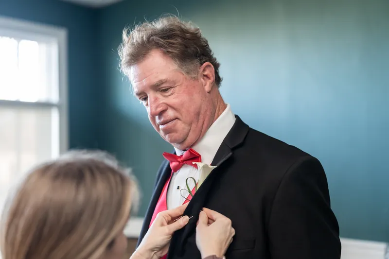 Bill adjusts his red bow tie while a woman helps him get ready in a teal room at The White Room.