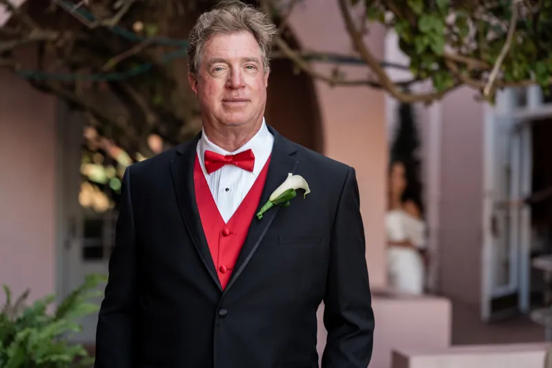 Bill smiles at the camera wearing a black suit, red vest, and red bow tie with a white boutonniere.