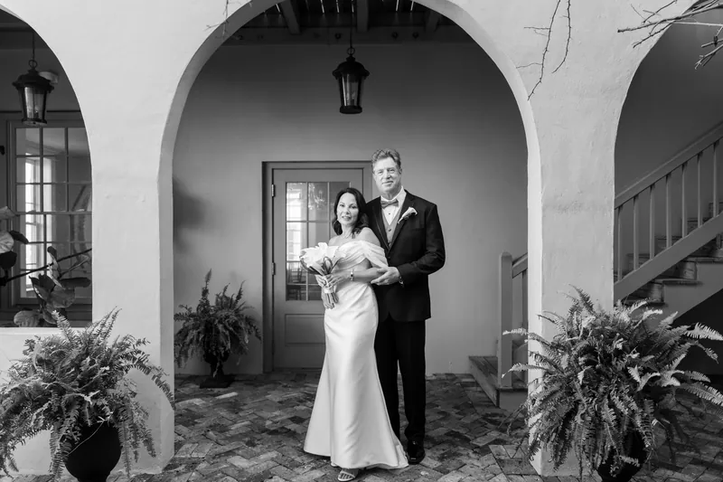 Rose Ann and Bill stand under a white arched doorway at The White Room, she in a flowing white gown and he in a dark suit with boutonniere.
