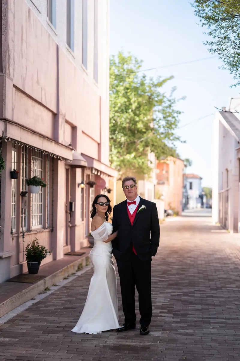Rose Ann and Bill walk down a brick pathway at The White Room, she holding her bouquet in a strapless white gown and he in a red-accented black suit.