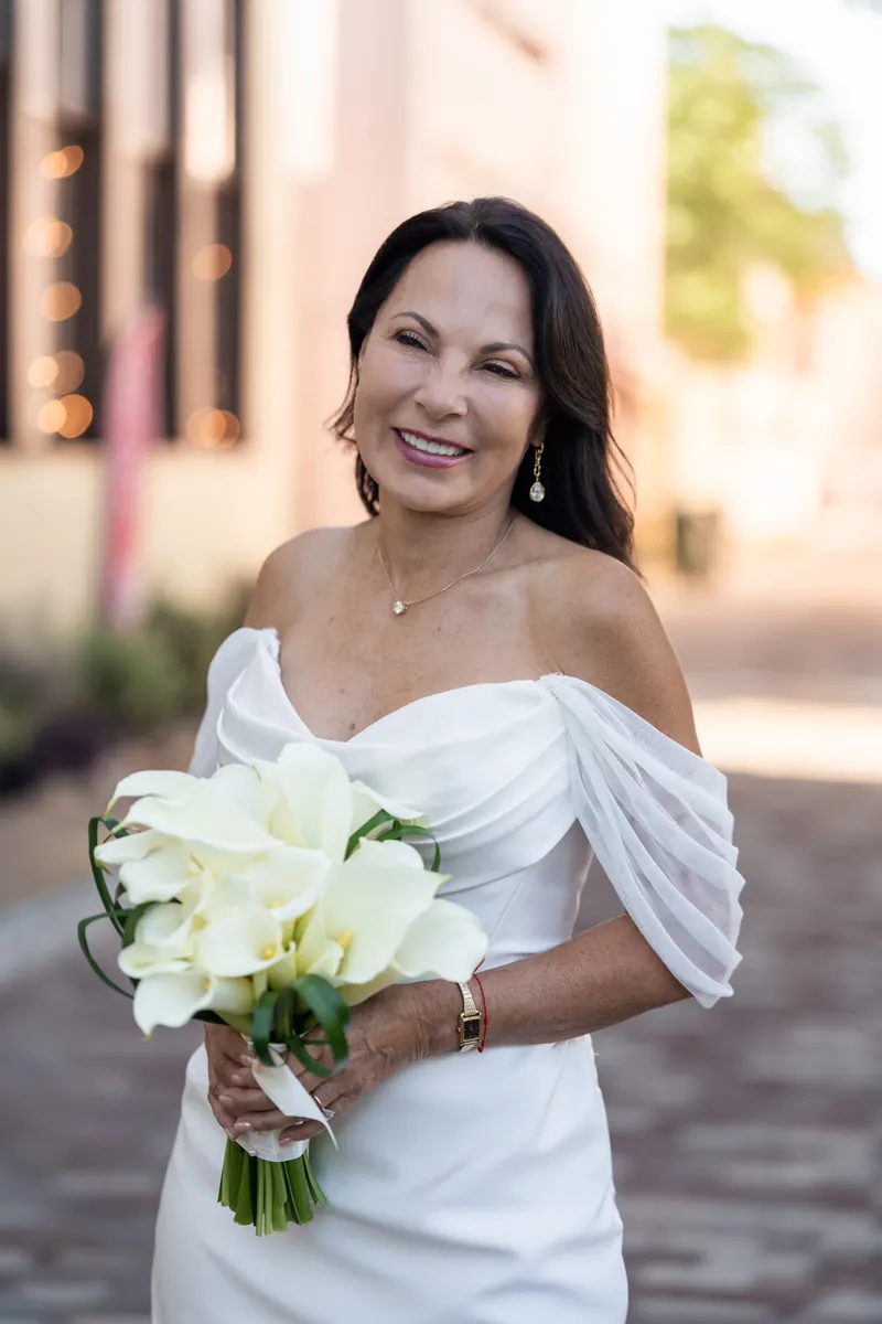 Rose Ann smiles at the camera holding a white orchid bouquet, wearing an off-shoulder white gown with a delicate necklace.