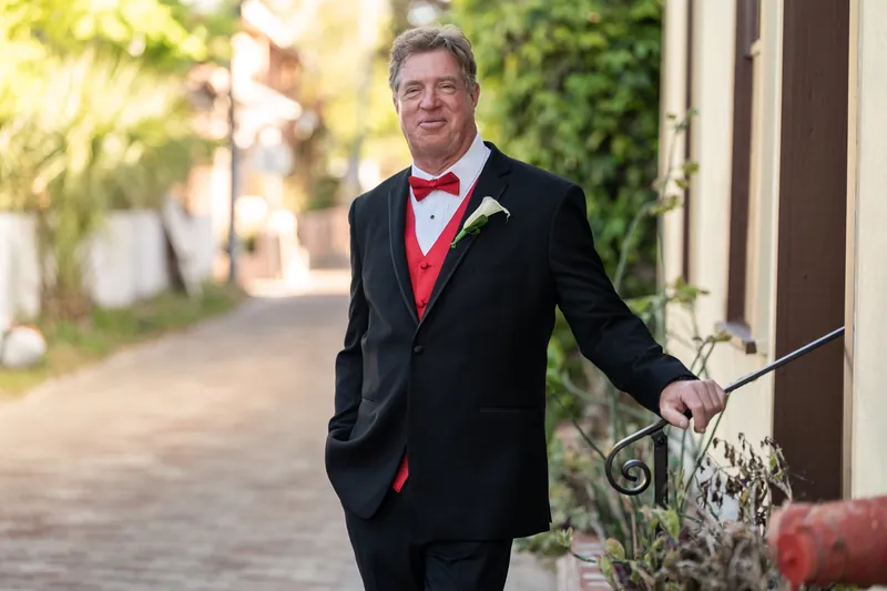 Bill walks down a shaded brick pathway at The White Room wearing a black suit with red vest and boutonniere.