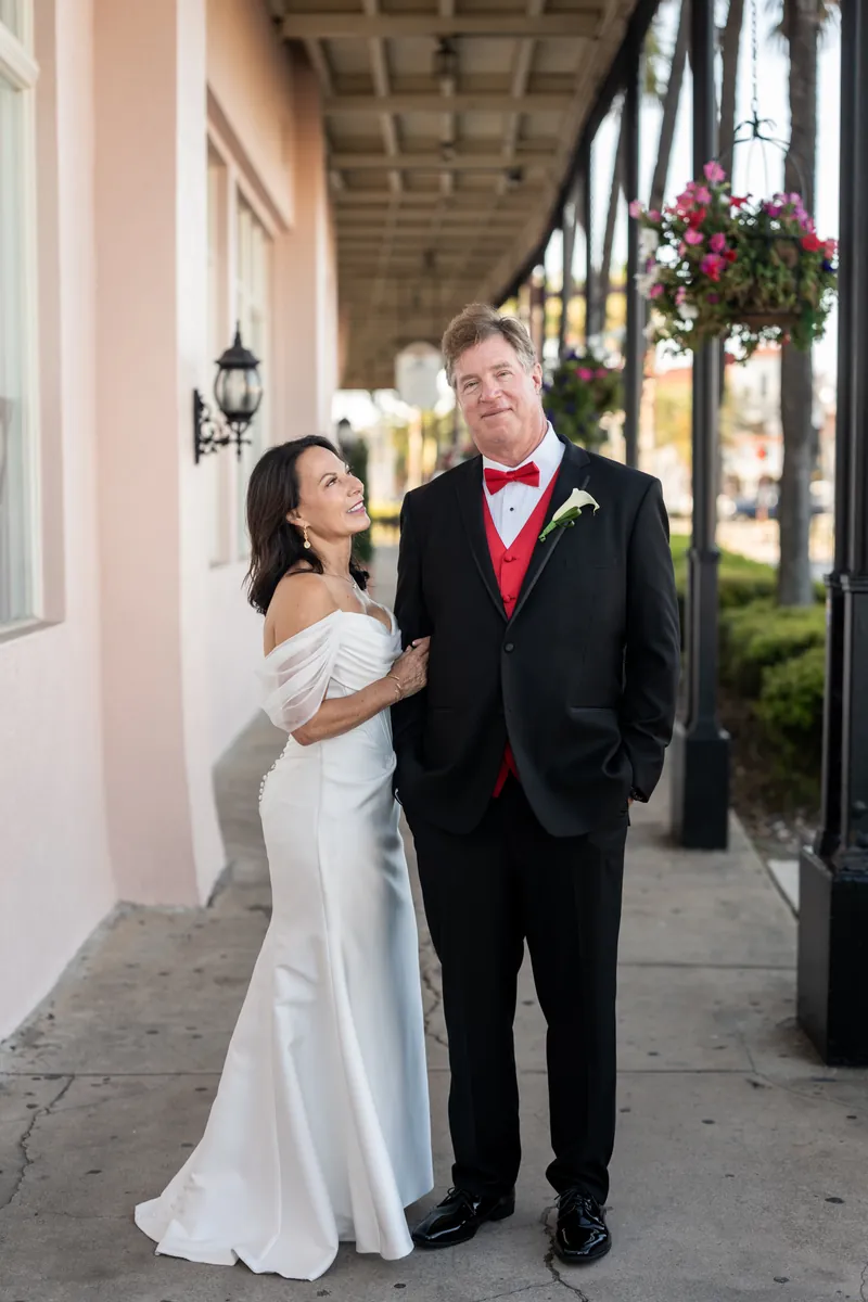Rose Ann gazes up at Bill as they stand together under The White Room's covered porch, she in white and he in black with red details.