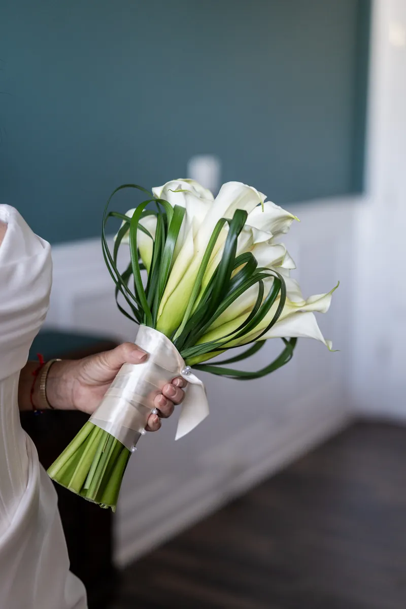 Bride's hand holding white calla lily bouquet with dark green ribbon at The White Room.