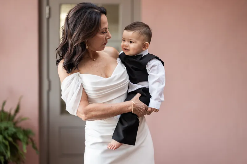Rose Ann in off-shoulder white dress holding a young boy in formal attire against pink wall.