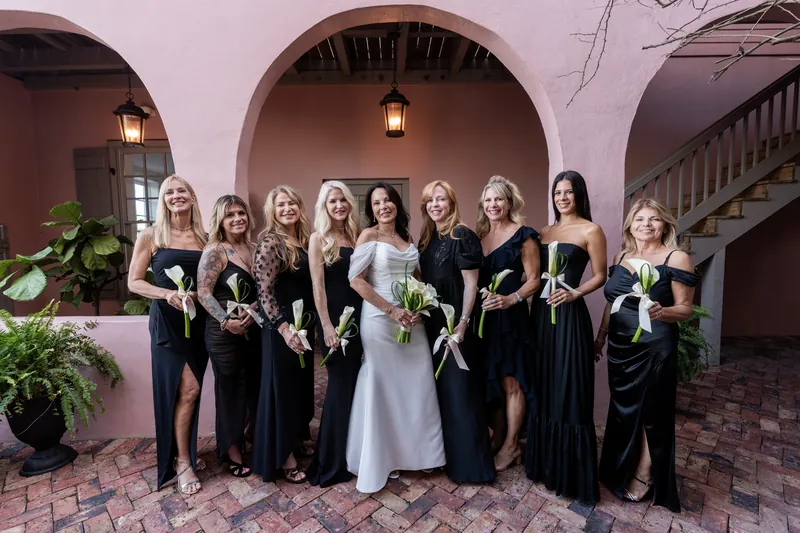 Rose Ann in white gown with nine bridesmaids in black dresses holding bouquets under arches at The White Room.