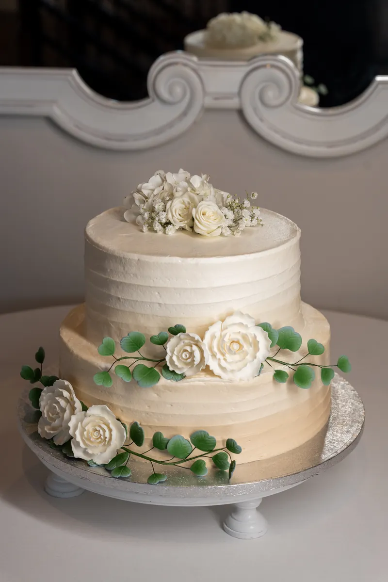Two-tier wedding cake with ivory frosting, white roses, and greenery on gray pedestal stand.