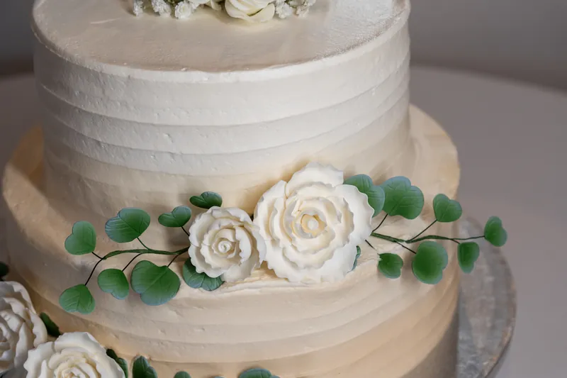 Two-tiered white wedding cake with textured frosting decorated with white fondant roses and green eucalyptus garland.