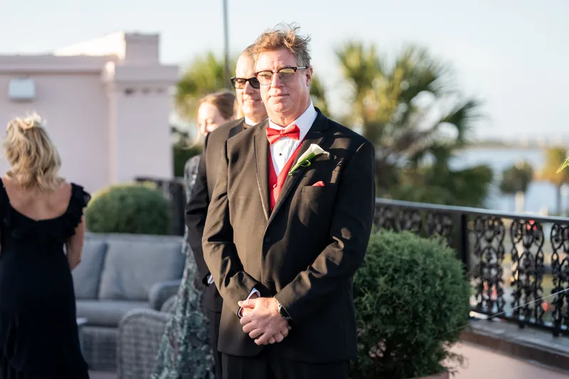 Bill stands at the altar wearing a dark suit with red boutonnière, smiling before the ceremony begins.