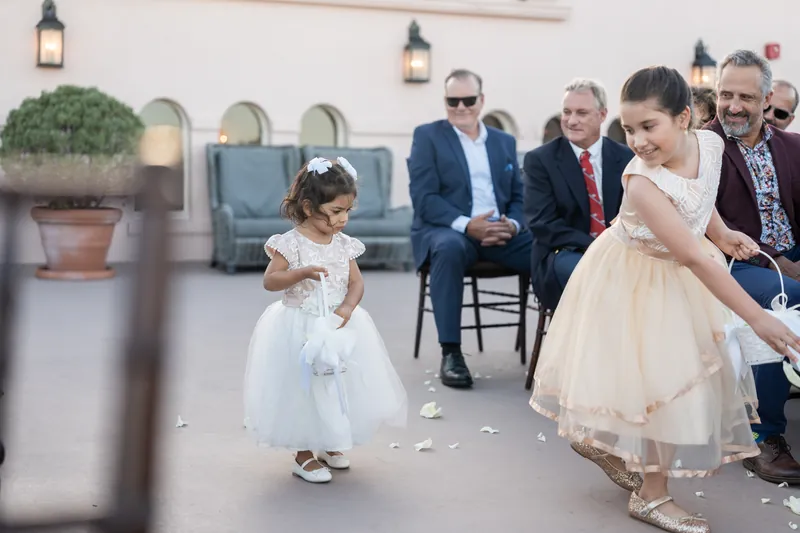 Young flower girl in white dress walks down the aisle as wedding guests seated behind her watch.