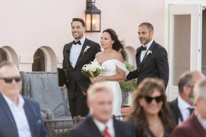 Rose Ann in white wedding dress holds bouquet while walking down aisle with Bill and groomsman at The White Room ceremony.