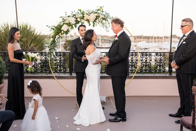 Bill kisses Rose Ann's hand during ceremony under white floral arch with city skyline visible behind them.