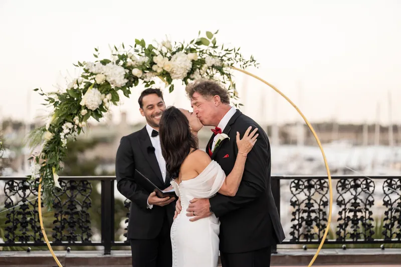 Bill embraces Rose Ann under gold arch after ceremony as officiant watches at The White Room rooftop venue.