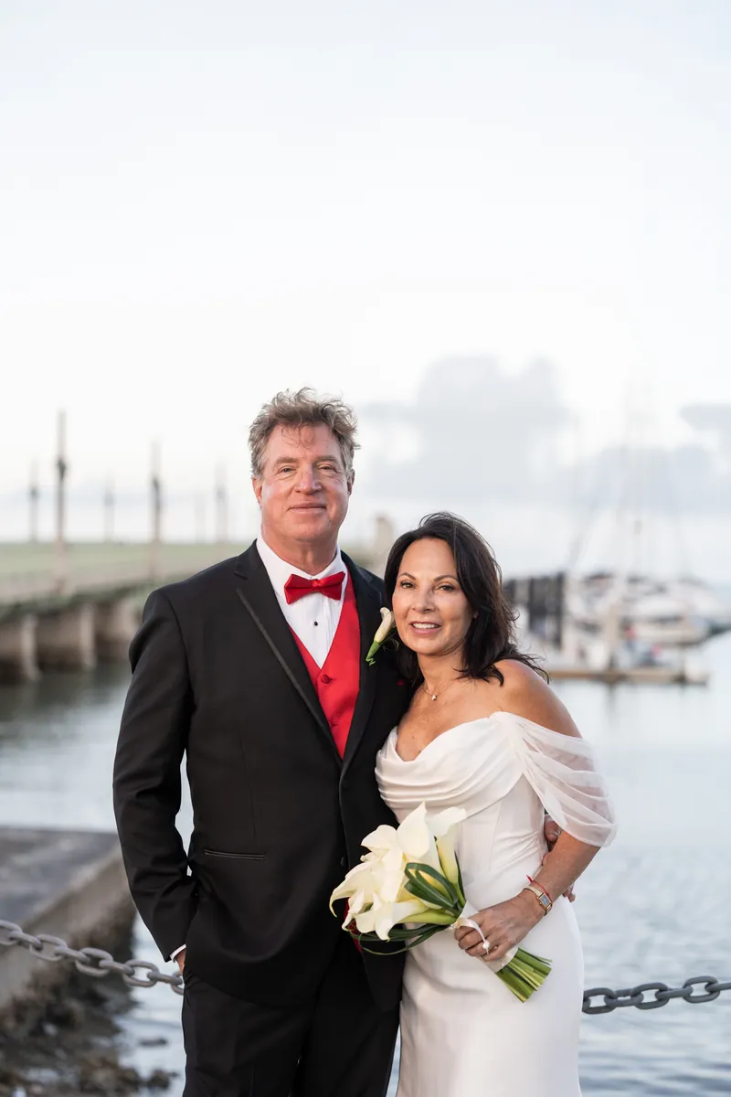 Bill wears a black tuxedo with red bow tie and Rose Ann holds white flowers in a cream off-shoulder wedding dress by the water.
