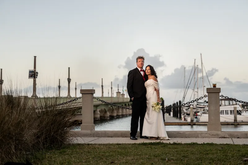Rose Ann in a white wedding dress and Bill in a black tuxedo stand together on a pier with sailboats and city skyline behind them.