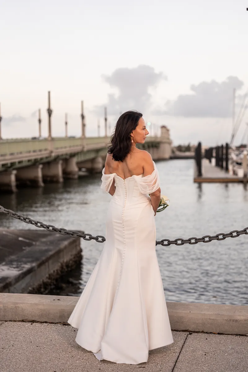 Rose Ann in an off-shoulder white wedding gown holds her bouquet while standing on a waterfront pier at The White Room.