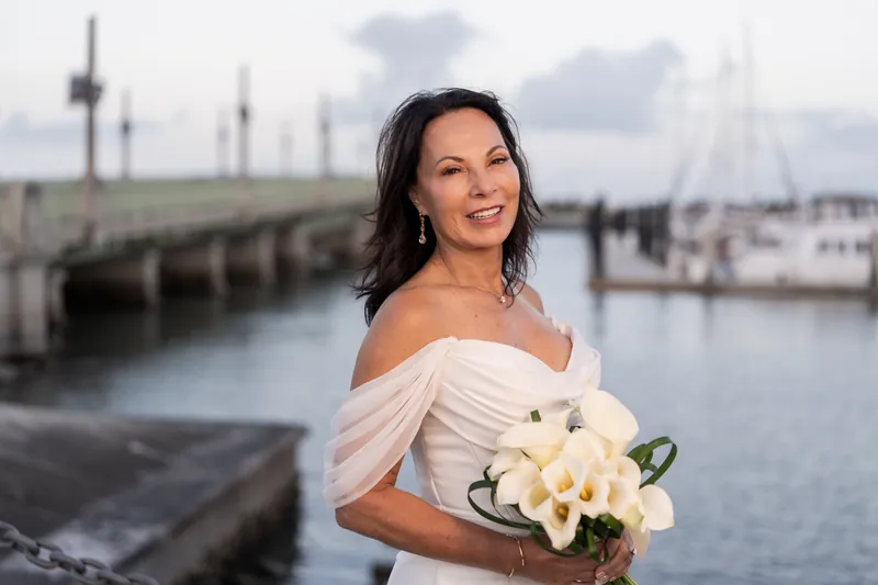 Rose Ann smiles while holding a white flower bouquet, wearing an off-shoulder white wedding dress with a waterfront backdrop.