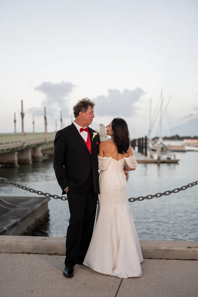 Bill and Rose Ann stand facing each other on a pier, Bill in black tuxedo with red bow tie, Rose Ann in white wedding dress.