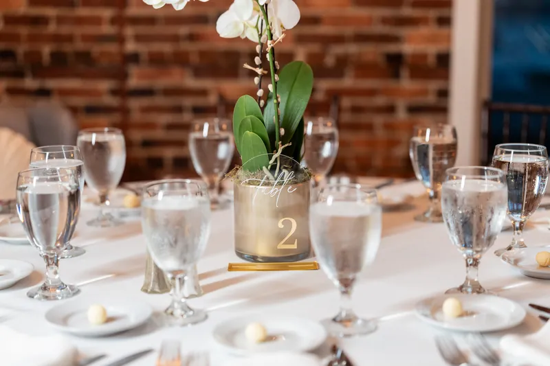 Dining table at The White Room set with water glasses, place settings, and white orchid centerpiece with gold table number two.