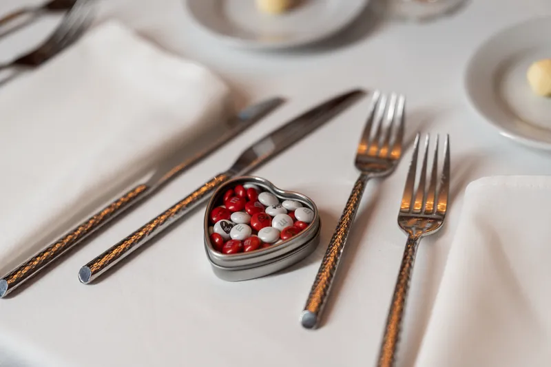 Gold-rimmed flatware and decorative heart-shaped box with red and white candies placed on white tablecloth at reception.