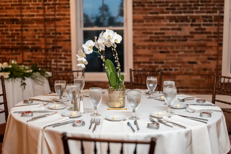 Round table with white linens, place settings, and tall white orchid arrangement in gold vase against exposed brick wall.