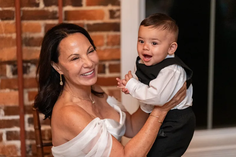 Woman in white off-shoulder dress holding smiling toddler in formal black and white outfit in front of brick wall.