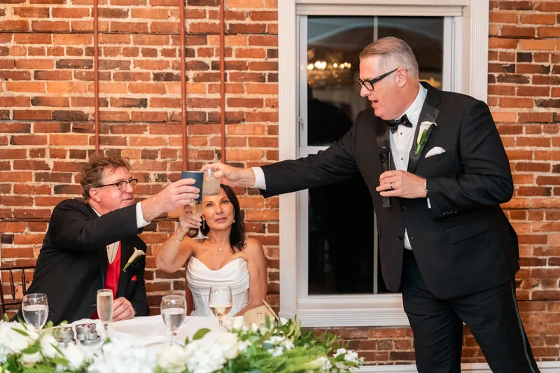A man in a tuxedo toasts with guests at a brick outdoor space during the wedding reception.