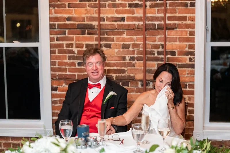Bill in a red vest and Rose Ann in a white dress laugh together at the reception table.
