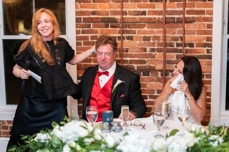 A woman in black speaks while Bill in a red vest and Rose Ann in white listen at the brick reception venue.