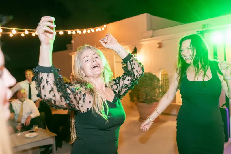 A blonde woman in a patterned jacket dances with her arms raised next to a woman in a green dress at the reception.