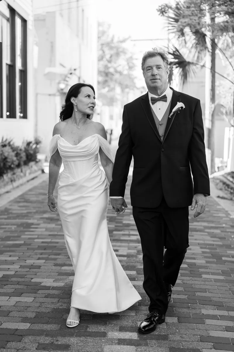 Rose Ann and Bill walk hand-in-hand down a palm-lined brick pathway, both in formal black-and-white wedding attire.