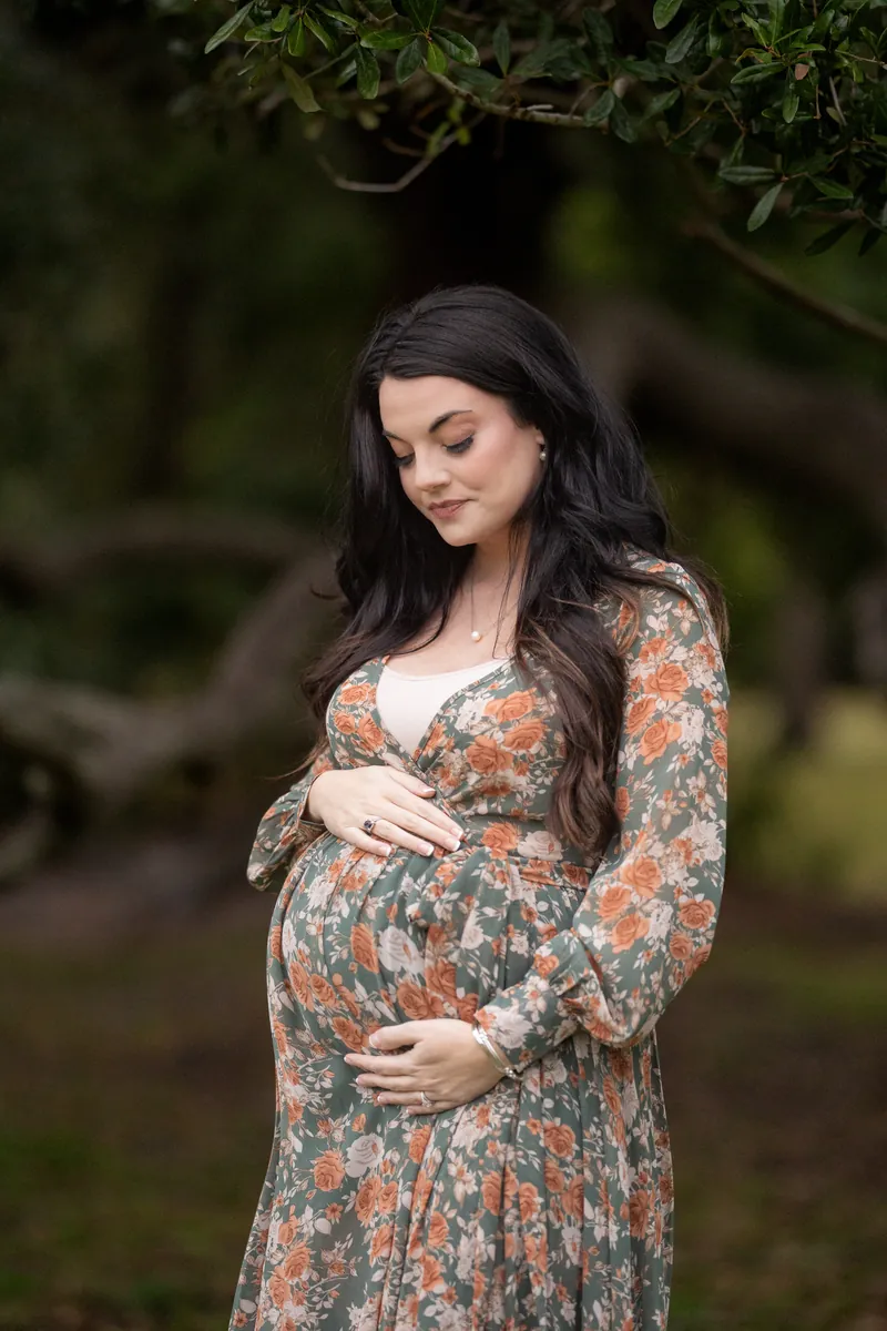 Valeen stands alone in a floral dress, gently holding her belly with both hands and gazing downward with a serene expression.