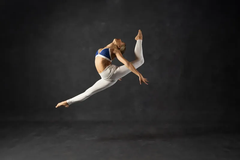 Sadie Dance executes a split jump in white leggings and navy crop top mid-leap against a dark backdrop.