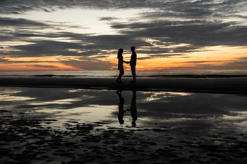 Sally and Pasha stand silhouetted holding hands on wet sand at sunset with dramatic sky and water reflections.