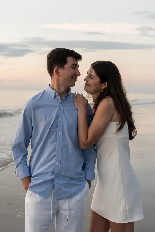 Pasha in light blue shirt and Sally in white dress face each other close on the beach at dusk.