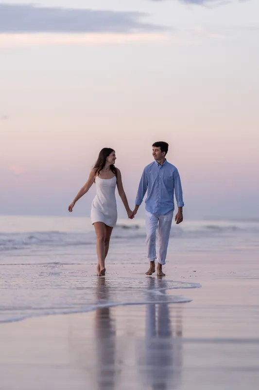 Sally and Pasha walk hand-in-hand through shallow water on the beach under a pink and purple sky.