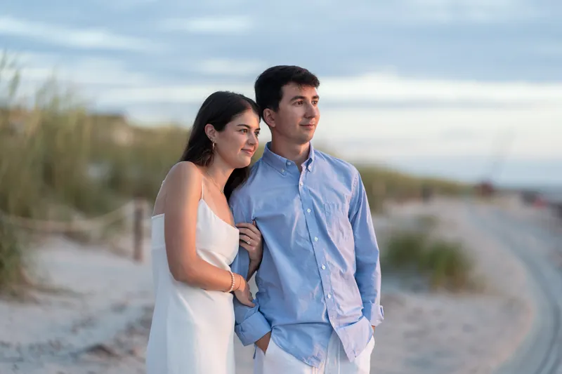 Sally in peach dress links her arm with Pasha in blue shirt as they stand on the beach near dune grass.