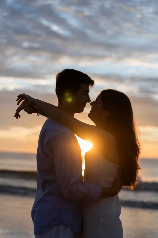 Sally and Pasha embrace at sunset on the beach with golden light between them and ocean waves behind.
